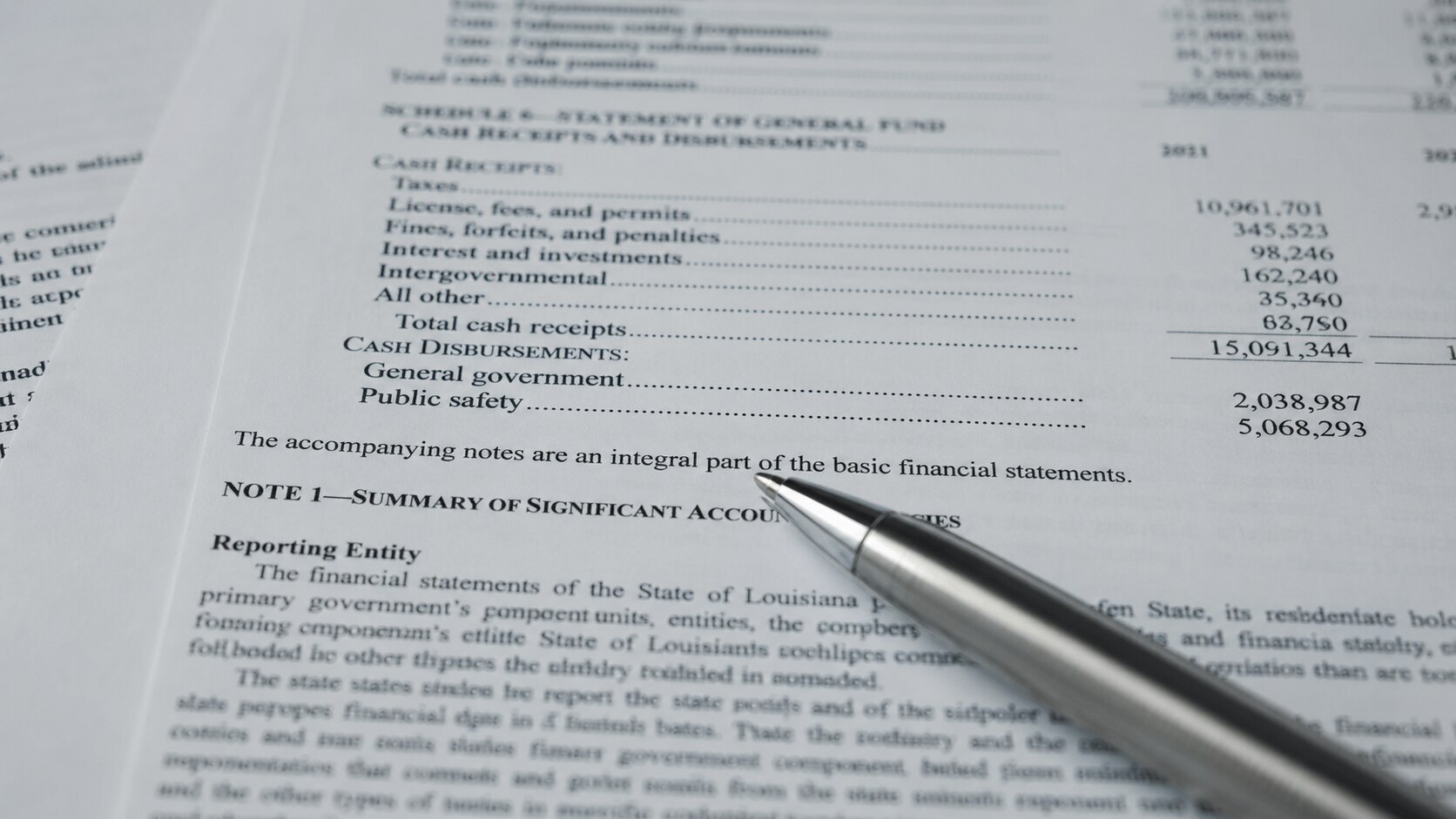 Close-up of a government financial statement showing cash receipts, disbursements, and accounting notes, with a silver ballpoint pen resting across the page — illustrating the kind of official document footnote in which the BEA disclosed its removal of investment silver from GDP trade data.