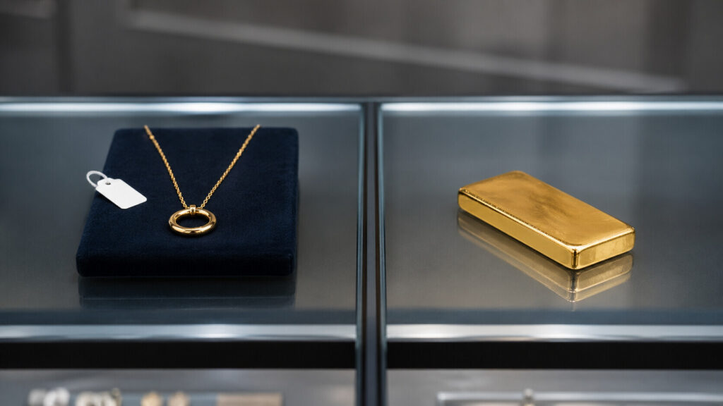 Gold necklace on a navy velvet pad with a price tag beside a plain gold bar on a glass jewelry counter — illustrating the difference between gold jewelry and gold bullion