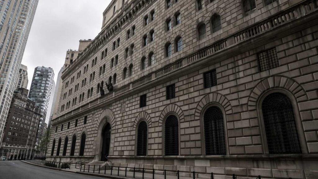 Exterior of the Federal Reserve Bank of New York building on Liberty Street, Manhattan, showing the neoclassical stone facade, arched windows, and surrounding skyscrapers under an overcast sky.