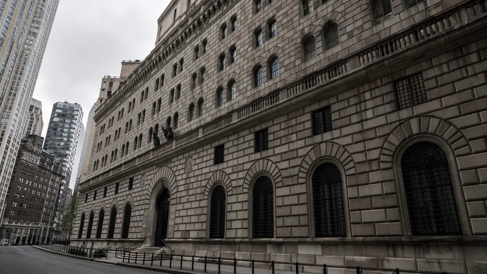 Exterior of the Federal Reserve Bank of New York building on Liberty Street, Manhattan, showing the neoclassical stone facade, arched windows, and surrounding skyscrapers under an overcast sky.