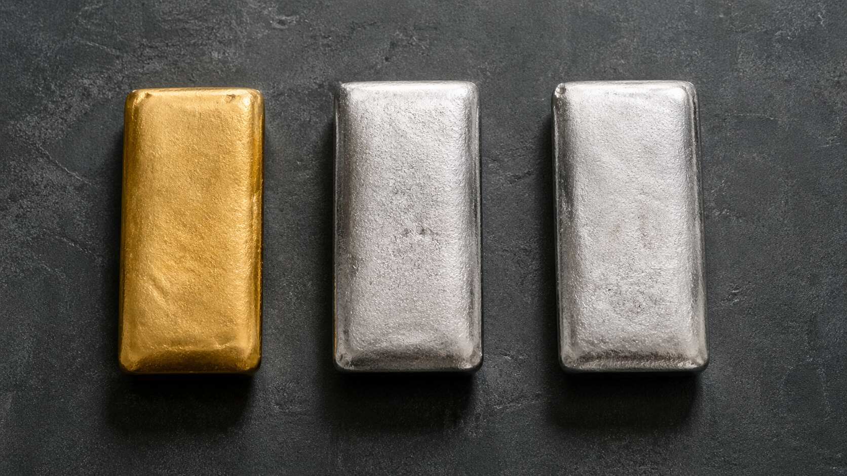 One gold bar and two silver bars on a dark stone surface