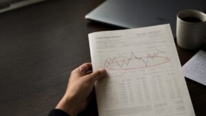 `Hand holding a financial research report with a red circle marking a key price range on a chart, dark walnut desk, coffee mug in background`