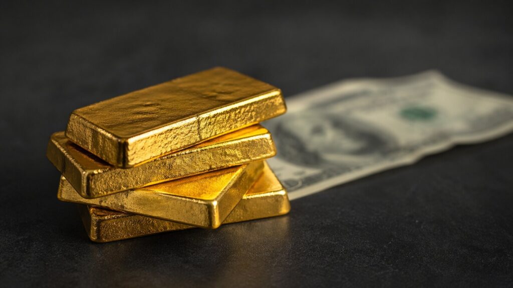 Gold bullion bars stacked on a dark surface with a U.S. hundred-dollar bill fading out of focus in the background