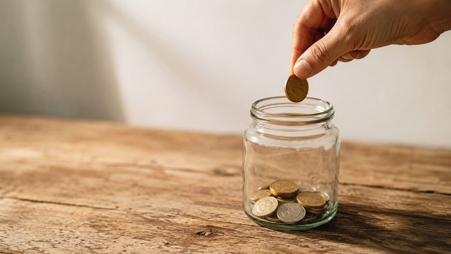 A hand dropping a gold coin into a glass jar containing gold and silver coins, illustrating dollar-cost averaging into precious metals