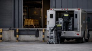 Two security personnel loading sealed transit cases into an armoured truck at a gold reserve warehouse loading bay, illustrating gold physical demand as metal moves out of Western storage facilities