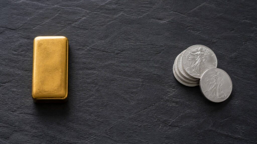 Gold bar and silver coins side by side on dark slate — illustrating the different portfolio roles of gold and silver as investment assets