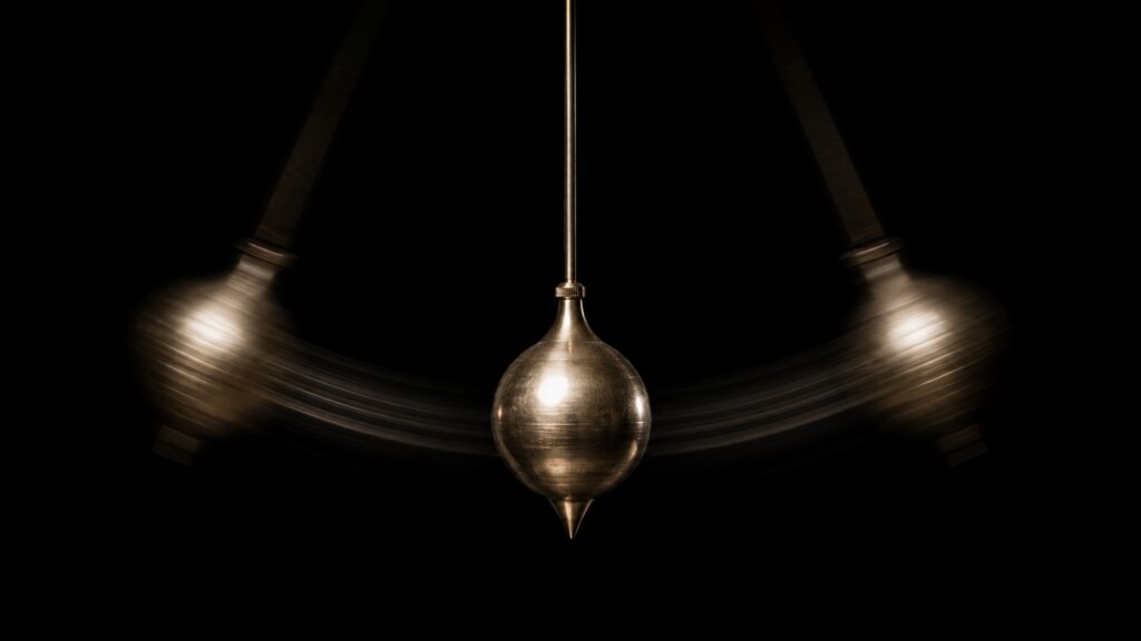 A brass pendulum bob centered at the midpoint of its swing, with symmetrical motion blur arcing to both sides against a black background — a visual metaphor for gold price cycles oscillating between extremes.
