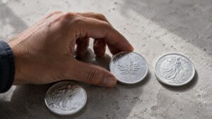 Silver Eagle, Maple Leaf, and Britannia coins on concrete with a hand mid-selection — silver eagle vs maple leaf vs britannia