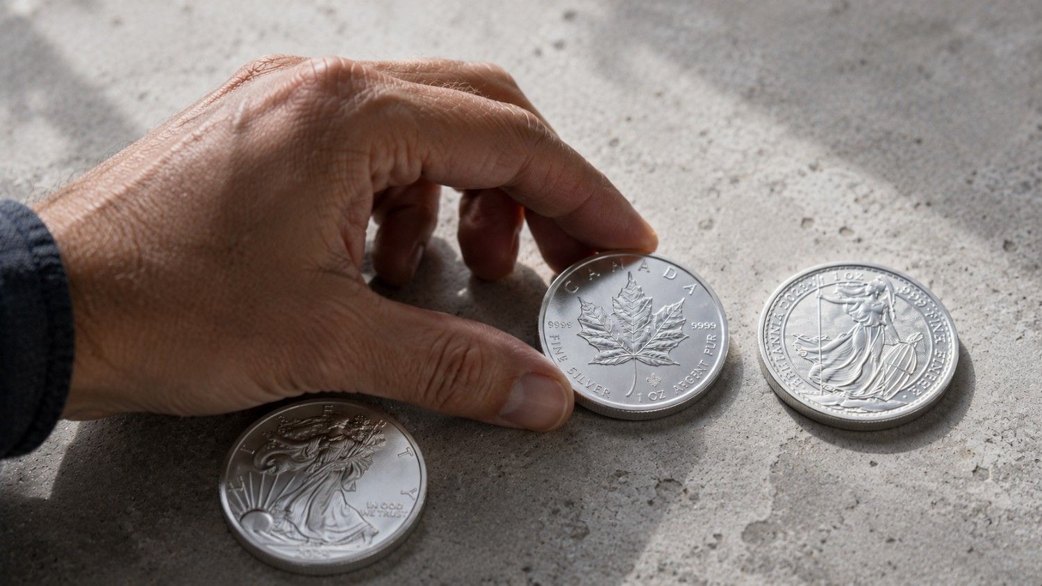 Silver Eagle, Maple Leaf, and Britannia coins on concrete with a hand mid-selection — silver eagle vs maple leaf vs britannia