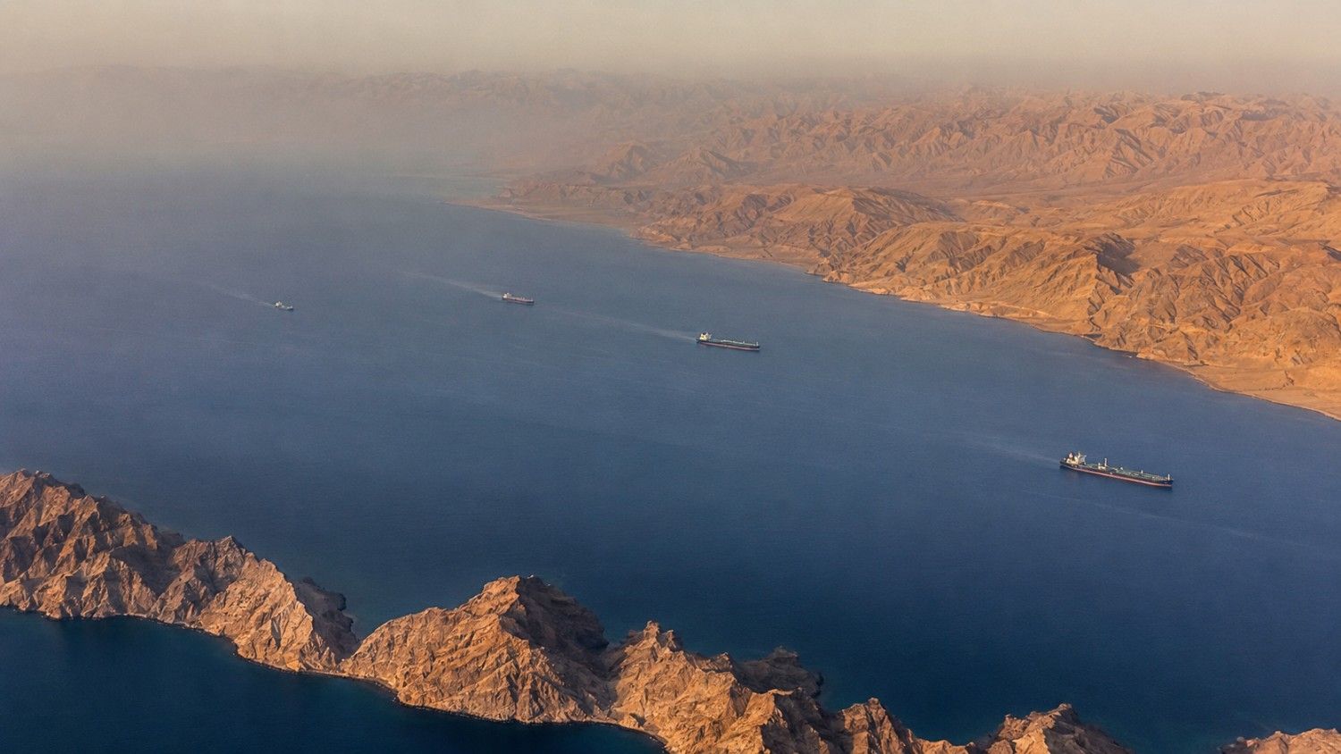 Aerial view of oil tankers transiting the Strait of Hormuz shipping lanes between the rocky coastlines of Iran and Oman, illustrating why gold is falling during a war that has disrupted global energy supply