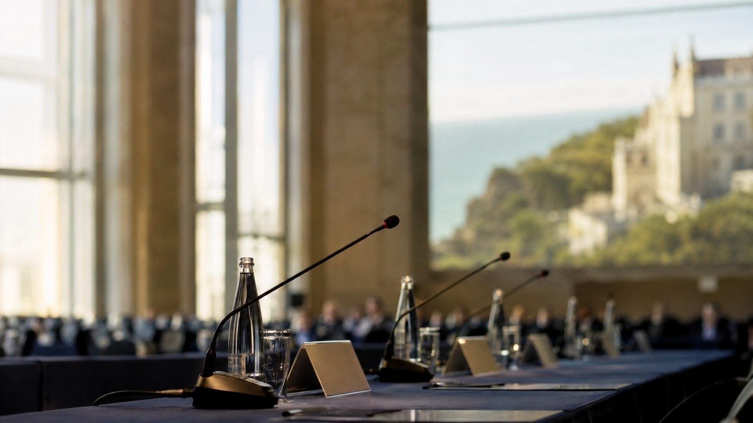 Empty conference microphones and name placards at a European central banking forum, with a coastal town in the background — why is gold up today amid Fed Chair Warsh's Sintra remarks