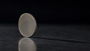 A plain metal coin balanced on its edge on dark stone, representing the coin-flip odds in the Waller rate hike gold price debate.
