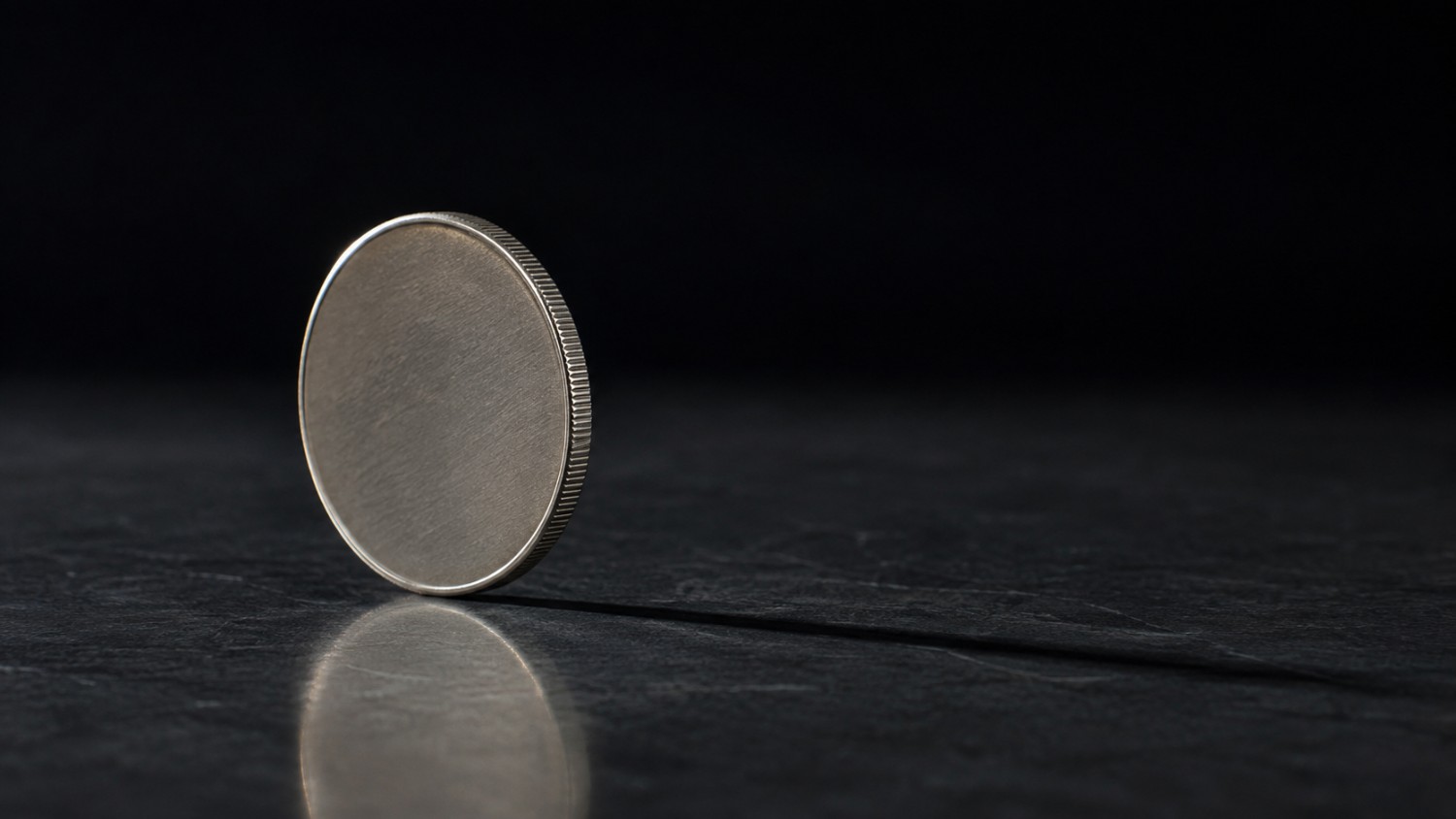 A plain metal coin balanced on its edge on dark stone, representing the coin-flip odds in the Waller rate hike gold price debate.